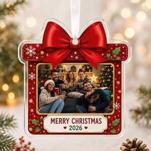 May include: A square-shaped Christmas ornament featuring a red gift box design with a large red bow at the top. The center displays a photograph of four people sitting together. The bottom includes the text Merry Christmas 2026 with two hearts.