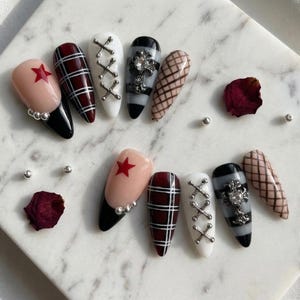May include: A set of ten long, almond-shaped artificial nails. Designs include a red star on a nude base, a black and white plaid pattern, white with silver cross accents, and a black and gray design with a silver embellishment.