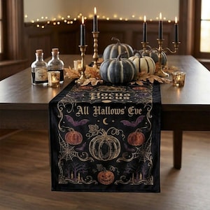 May include: A black velvet table runner featuring gold and purple printed designs of pumpkins, spiderwebs, bats, and trees. The text reads Hocus Pocus and All Hallows' Eve. The runner is decorated with real pumpkins and lit candles in gold holders.