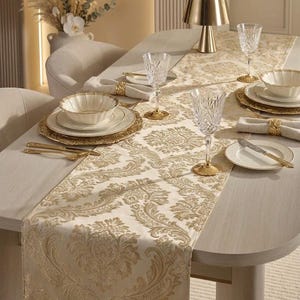 May include: A cream-colored table runner featuring a repeating gold damask pattern. The runner is set on a dining table with white plates, fluted bowls, crystal glasses, gold-toned cutlery, and gold napkin rings, creating a coordinated table setting.