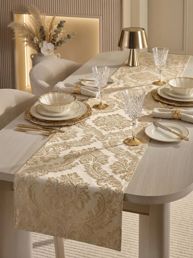 May include: A cream-colored table runner featuring a repeating gold damask pattern. The runner is set on a dining table with white plates, fluted bowls, crystal glasses, gold-toned cutlery, and gold napkin rings, creating a coordinated table setting.
