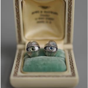May include: Two silver-coloured eye-shaped earrings, each with a detailed iris and pupil, displayed in a vintage jewellery box with a green velvet interior. The earrings have a unique, artistic design.