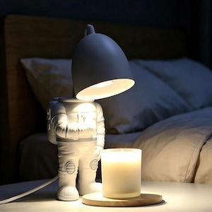 May include: A white astronaut-shaped table lamp with a dome-shaped shade emitting light downward, positioned next to a cylindrical white candle in a glass jar resting on a circular wooden base, creating a warm and focused illumination.
