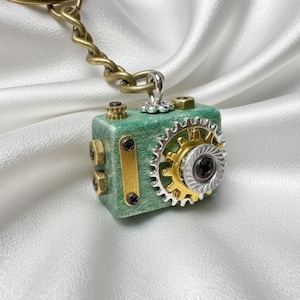 May include: A steampunk-style keychain featuring a small, green, cube-shaped charm. The charm has gold and silver gears, bolts, and accents. The keychain has a brass-colored chain and clasp, and is set against a white, silky background.