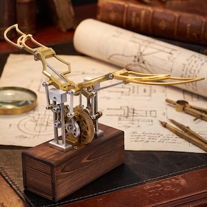 May include: A mechanical kinetic sculpture featuring a brass-colored bird-like wing mechanism mounted on a rectangular dark wood base. The assembly includes silver-toned metal pistons, vertical support rods, and a central gear system.