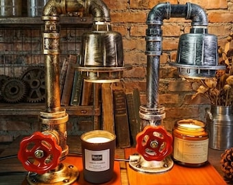 May include: Two industrial-style table lamps with a pipe design, featuring red valve handles and metallic shades. Each lamp sits on a wooden base, accompanied by candles. The background includes books and a brick wall.
