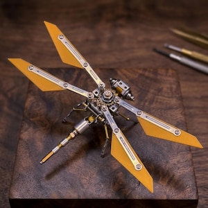 May include: A detailed steampunk dragonfly sculpture with yellow wings and intricate metalwork. The dragonfly is made of silver, gold, and brass-colored metal, with gears and rivets visible. The wings are a bright yellow color. The sculpture is displayed on a dark wooden surface.