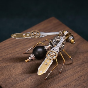 May include: A steampunk-style insect sculpture made of metal components, including gears, screws, and brass accents. The insect has a black spherical body and detailed wings, legs, and antennae. It is displayed on a wooden surface.