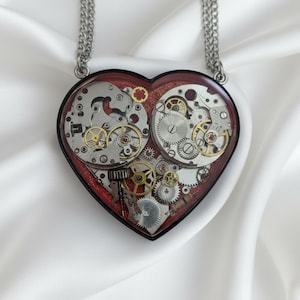May include: Heart-shaped pendant necklace featuring watch gears and cogs encased in resin. The pendant has a black border and a red glitter background. The silver-toned chain is visible above the pendant.