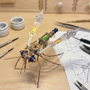 Puede incluir: Escultura de libélula de estilo steampunk hecha de engranajes metálicos, tubos y otros componentes mecánicos. Las alas son plateadas y doradas, y el cuerpo presenta detalles azules y dorados. La escultura está sobre una superficie de madera.