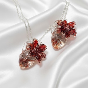 May include: Two clear glass heart pendants with red floral accents hang from silver chains. The pendants are displayed on a white satin background, showcasing the intricate details of the jewelry.