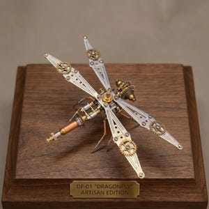 Puede incluir: Una escultura de libélula de estilo steampunk hecha de componentes metálicos, incluyendo engranajes y tubos. La libélula está montada sobre una base de madera con una placa de latón que dice "DF-01 'DRAGONFLY' ARTISAN EDITION". Las alas son plateadas.