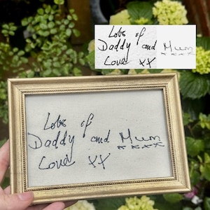 May include: A framed artwork with a gold-coloured frame. The piece displays handwritten text in black ink: "Lots of Daddy and Mum Loved XX". The background is a light beige fabric, creating a simple, heartfelt design.