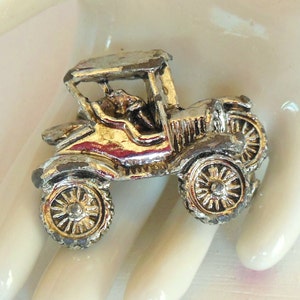 May include: Vintage-style car brooch with a silver and gold finish. The brooch features a detailed design of an antique car with spoked wheels and a convertible top. A classic accessory for collectors.