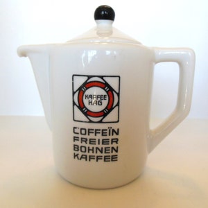 May include: White ceramic coffee pot with a lid and handle. The pot features a black and red logo that says "KAFFEE HAG" and the words "COFFEIN FREIER BOHNEN KAFFEE" in black text. The lid has a black knob.