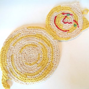 May include: A handmade, crocheted cat-shaped trivet in yellow and cream colors. The trivet features a spiral design on the body and a cat face with red and green details. The trivet is designed to protect surfaces from hot items.