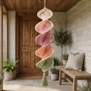 May include: A hanging spiral wind spinner crafted from crochet yarn in a gradient of cream, soft pink, dusty rose, peach, and sage green. The structure features an openwork stitch pattern and terminates in a single wooden bead and a sage green tassel.