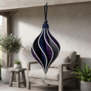 Puede incluir: Un adorno de ganchillo colgante con forma de lágrima y paneles en espiral en negro, azul marino, morado y gris. Los bordes están ribeteados con hilo plateado y un pequeño dije de media luna plateado cuelga de la punta inferior.
