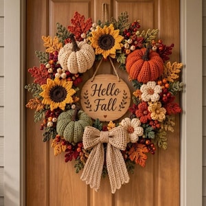 Op de afbeelding: Een ronde gehaakte krans met oranje, groene en crèmekleurige pompoenen, gele zonnebloemen en herfstkleurige bladeren. Een centraal houten bordje toont de tekst Hello Fall met een klein hartje. Onderaan hangt een grote beige gehaakte strik.