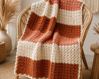 Puede incluir: Una manta de ganchillo con anchas rayas horizontales en tonos alternos de terracota, naranja óxido y crema. La manta presenta un patrón de puntos texturizado y repetitivo, con un borde festoneado de color crema en la parte inferior.