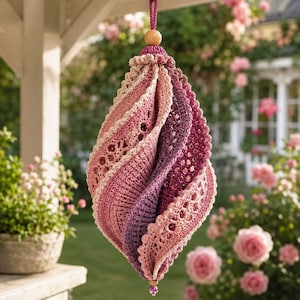 May include: A hanging crochet ornament featuring a spiralling, multi-layered design in shades of pink and purple. The piece is constructed with intricate lace-like stitching and scalloped edges, suspended by a cord with a wooden bead at the top.