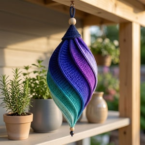 May include: A hanging crochet ornament featuring a spiraling, teardrop-shaped design. The textile transitions in color from deep purple at the top, through shades of blue, to a light teal at the bottom. It is accented with wooden beads and a dark cord.