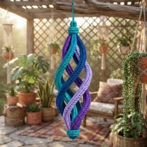 Puede incluir: Un adorno en espiral colgante hecho de hilo de ganchillo en un degradado de colores que incluye morado oscuro, azul real, turquesa y lavanda claro. La estructura presenta un diseño central retorcido con un lazo anudado en la parte superior e inferior.
