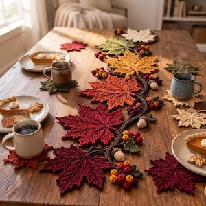Pode incluir: Um caminho de mesa em crochê com uma vinha central de folhas de bordo em tons outonais de vermelho, laranja, amarelo e verde, acentuado com pequenas bagas e bolotas em crochê, acompanhado por bases para copos em forma de folha.
