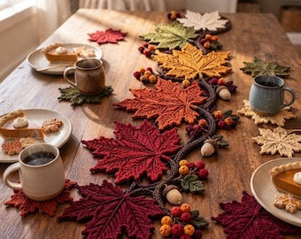 Puede incluir: Un camino de mesa de ganchillo con una enredadera central de hojas de arce en colores otoñales, tonos rojos, naranjas, amarillos y verdes, decorado con pequeñas bayas y bellotas tejidas, junto con posavasos a juego en forma de hoja.
