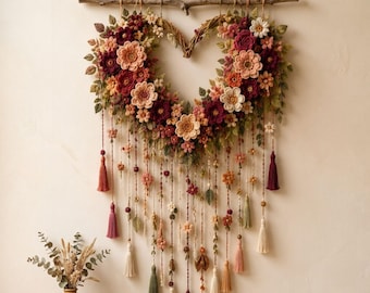 Puede incluir: Un adorno de pared en forma de corazón con flores de ganchillo en tonos burdeos, melocotón y crema, acentuado con hojas verdes. El arreglo cuelga de una rama de madera e incluye múltiples hilos colgantes adornados con borlas.