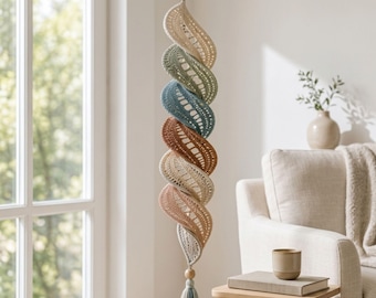Puede incluir: Una decoración colgante en espiral hecha de tela de ganchillo en una secuencia vertical de segmentos color crema, verde salvia, azul verdoso, terracota, crema y rosa empolvado. La pieza tiene una cuenta de madera arriba y abajo, terminando en una borla azul claro.