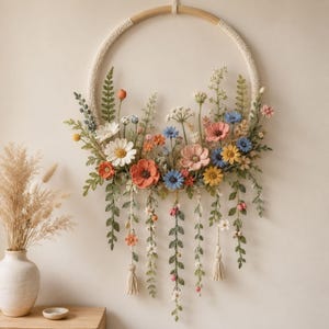 Op de afbeelding: Een ronde wandhanger met een houten ring omwikkeld met crèmekleurig garen, versierd met diverse gehaakte wilde bloemen, groene bladeren en varens in tinten oranje, blauw, roze, geel en wit, met naar beneden hangende bloemranken.