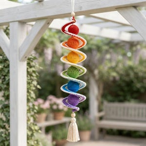 Puede incluir: Una decoración colgante en espiral con seis esferas de ganchillo en rojo, naranja, amarillo, verde, azul y morado. Las esferas están unidas por una estructura en espiral de ganchillo blanca, rematada con una cuenta de madera y una borla.