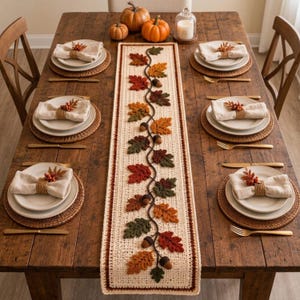 May include: A cream-colored crochet table runner featuring a central vine pattern with textured autumn leaves in shades of orange, red, brown, and green, accented with small brown acorns, laid on a wooden dining table with four place settings.