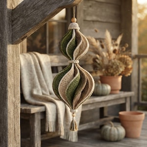 Puede incluir: Un adorno de ganchillo colgante con forma de doble bulbo en espiral, compuesto por segmentos alternos de hilo verde oliva y beige. La pieza está decorada con pequeñas cuentas de madera en la parte superior e inferior y un borla de hilo.