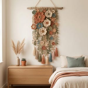 May include: A wall hanging featuring a collection of crocheted flowers in shades of terracotta, sage green, cream, and dusty blue. The floral arrangement is suspended from a natural wooden branch with hanging yarn tassels and wooden beads.