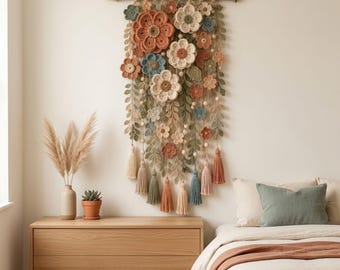 Puede incluir: Un tapiz de pared con una colección de flores de ganchillo en tonos terracota, verde salvia, crema y azul polvoriento. El arreglo floral cuelga de una rama de madera natural con borlas de hilo colgantes y cuentas de madera.