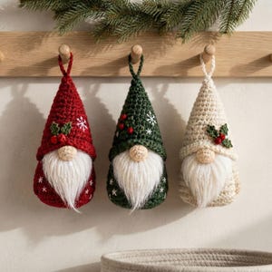Puede incluir: Tres adornos de gnomo de ganchillo colgados de un perchero de madera. El gnomo de la izquierda es rojo con copos de nieve blancos, el central es verde oscuro con copos de nieve blancos y el de la derecha es color crema. Cada uno tiene barba blanca.