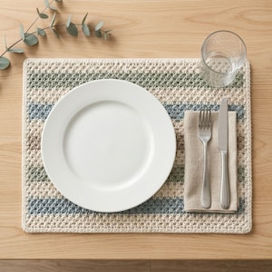 May include: A rectangular crochet placemat featuring horizontal stripes in cream, sage green, and dusty blue. A plain white ceramic dinner plate sits in the center, accompanied by a beige linen napkin, a stainless steel fork, and a knife.