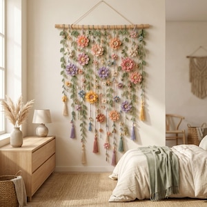 May include: A wall hanging featuring a horizontal wooden rod with multiple vertical strands of crocheted green leaves and various colorful flowers in shades of pink, purple, orange, and yellow, finished with dangling tassels at the bottom.