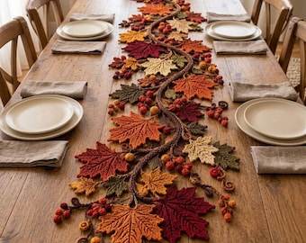 Puede incluir: Un camino de mesa decorativo que presenta una larga enredadera sinuosa adornada con hojas de arce tejidas a ganchillo en tonos rojo, naranja, amarillo y verde, intercaladas con pequeños racimos de bayas redondas en colores otoñales.