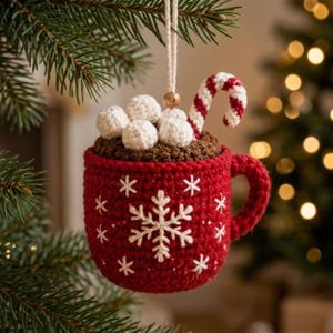 May include: A red crocheted mug ornament featuring a white embroidered snowflake pattern, filled with brown crocheted hot cocoa topped with white crocheted marshmallows and a red and white striped crocheted candy cane, hanging by a string.