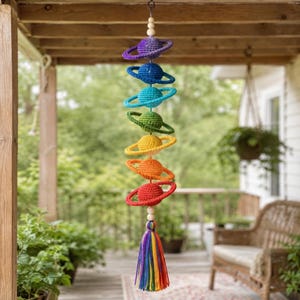 May include: A vertical hanging mobile featuring seven crocheted spheres with rings, arranged in a spectrum of colors from purple to red. Each planet-like sphere is separated by small wooden beads, and the piece ends with a multi-colored tassel.