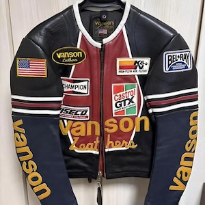 Peut inclure: Veste en cuir noire avec des détails rouges et blancs, ornée d'écussons avec du texte et des logos. Les manches sont bleues avec "Vanson" en lettres dorées. Un écusson du drapeau américain est visible sur la manche gauche. La veste est suspendue à un cintre noir.