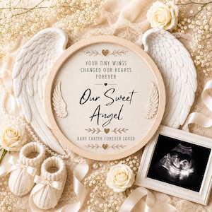 May include: A circular wooden frame containing a fabric insert with the text "YOUR TINY WINGS CHANGED OUR HEARTS FOREVER", "Our Sweet Angel", and "BABY CARTER FOREVER LOVED", flanked by two white angel wings and a baby ultrasound photo.