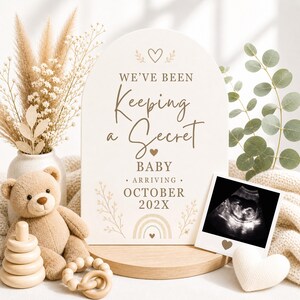 May include: An arched sign with the text "WE'VE BEEN Keeping a Secret BABY ARRIVING OCTOBER 202X" featuring a small rainbow and heart icons. Beside it sits a sonogram in a white frame, a tan teddy bear, a wooden stacking toy, and a wooden ring.