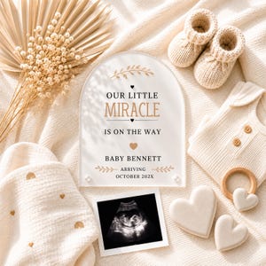 May include: An arched acrylic sign with the text OUR LITTLE MIRACLE IS ON THE WAY BABY BENNETT ARRIVING OCTOBER 202X, accompanied by a sonogram, beige knit baby booties, a ribbed onesie, a wooden ring, and two heart-shaped ornaments.