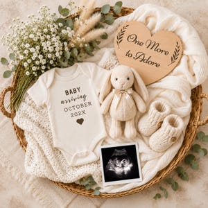Op de afbeelding: Een witte babyromper met de tekst "BABY arriving OCTOBER 202X" en een hartje, een crèmekleurig pluche konijn, gebreide babyslofjes, een hartvormig houten bordje met "One More to Adore", een echofoto en witte bloemen in een gevlochten mand.