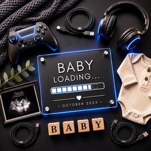 May include: A black acrylic sign with the text BABY LOADING... and a progress bar, featuring the date OCTOBER 202X. The sign is surrounded by a game controller, headphones, a baby bodysuit, an ultrasound photo, and wooden blocks spelling BABY.