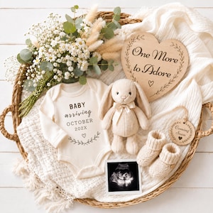 Op de afbeelding: Een crèmekleurige babyromper met de tekst Baby arriving October 202X, een beige pluche konijn, een paar gebreide babyslofjes, een hartvormig houten bordje met de tekst One More to Adore, een rond Coming Soon-label en een echofoto.